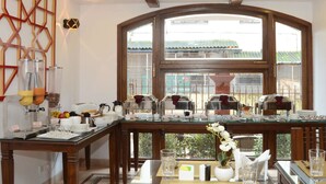 Disposição de buffet de café da manhã com diversos alimentos e bebidas, uma mesa de madeira e vista para o exterior através de uma grande janela.