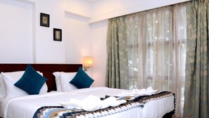 Quarto de hotel com cama grande, duas almofadas, cabeceira, luminária na parede e cortinas leves.
