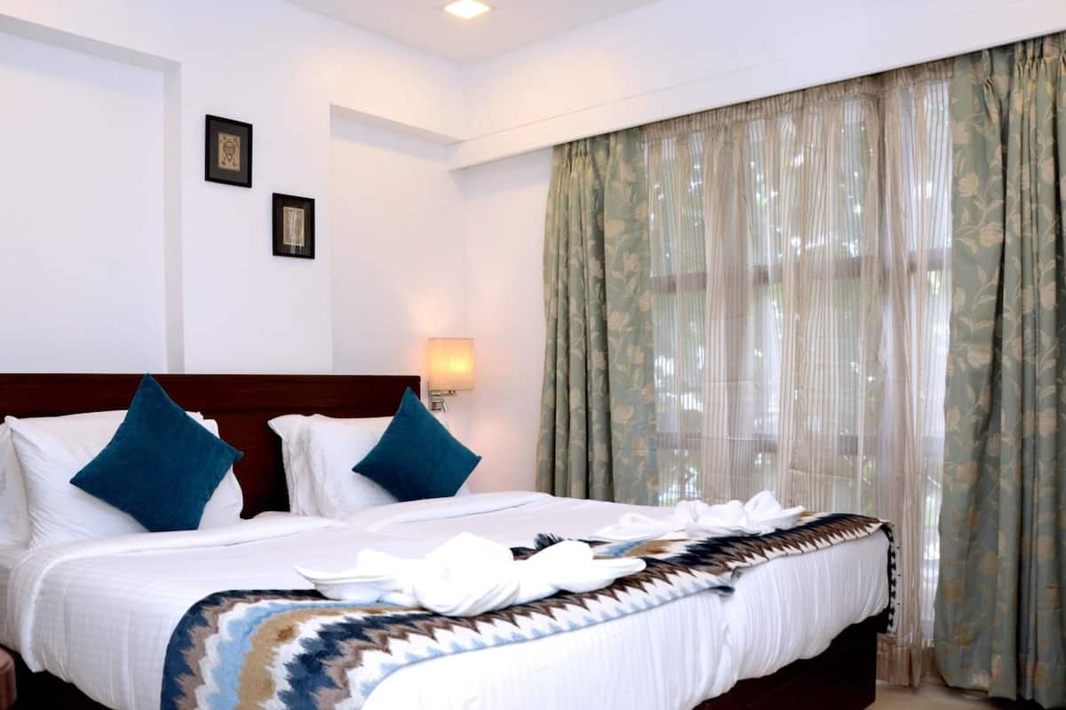 A hotel room with a large bed, two pillows, a headboard, a wall-mounted lamp, and sheer curtains.