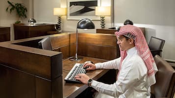 Un homme vêtu d’une chemise blanche et d’un couvre-chef traditionnel travaille à un bureau en bois devant un ordinateur, dans une pièce éclairée par une lampe de bureau et ornée d’œuvres d’art encadrées au mur.