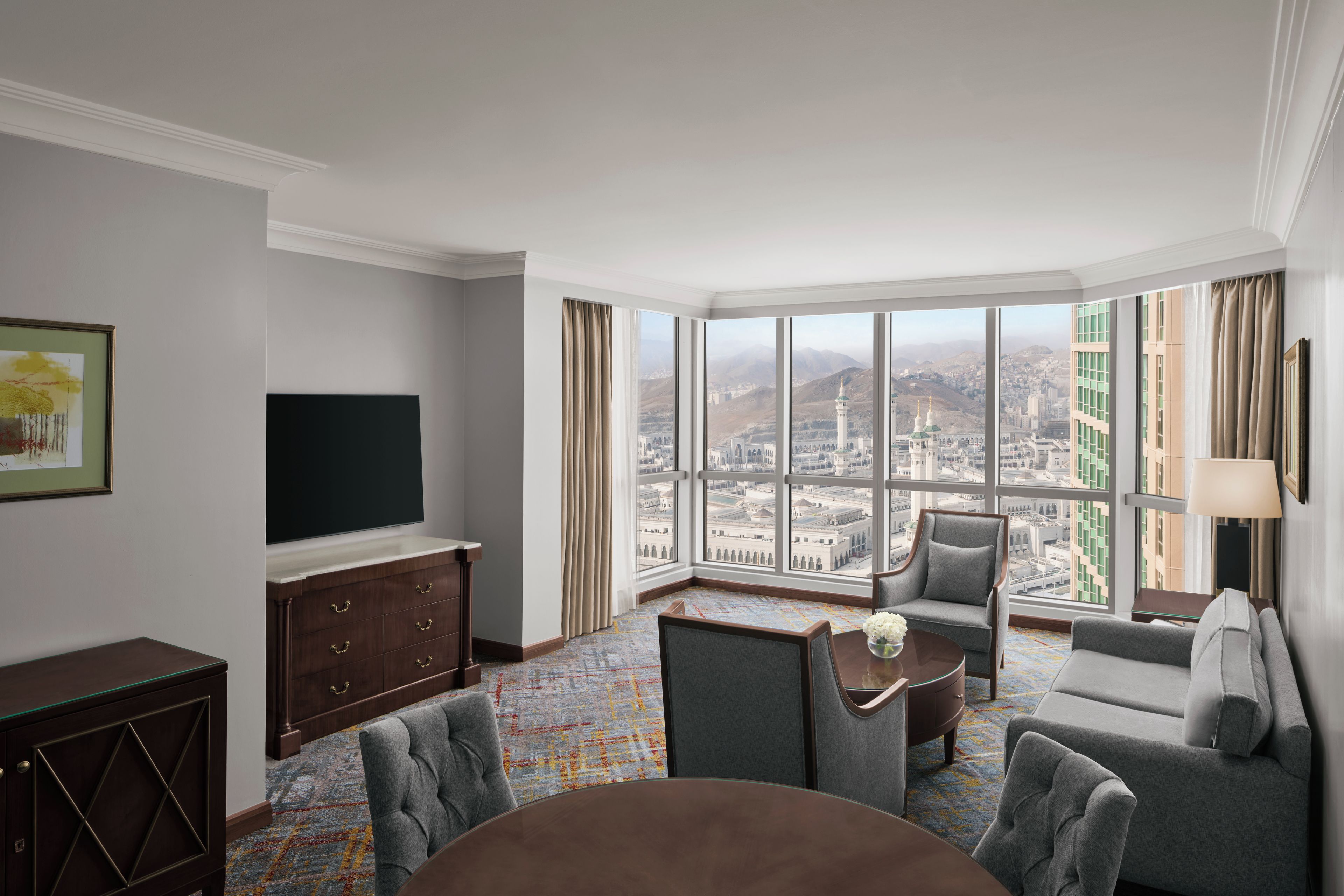 A hotel room with a large window offering a city view, a flat-screen TV, a sofa, a chair, and a small table.
