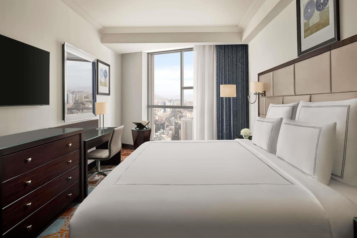 A hotel room with a large bed, a desk, a chair, a dresser, a TV, and a window with a city view.