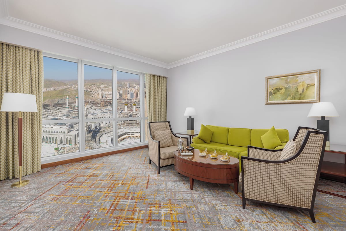 Een hotelkamer met een groot raam dat uitzicht biedt op de stad, een groene bank, twee fauteuils, een klein rond tafeltje en twee vloerlampen.
