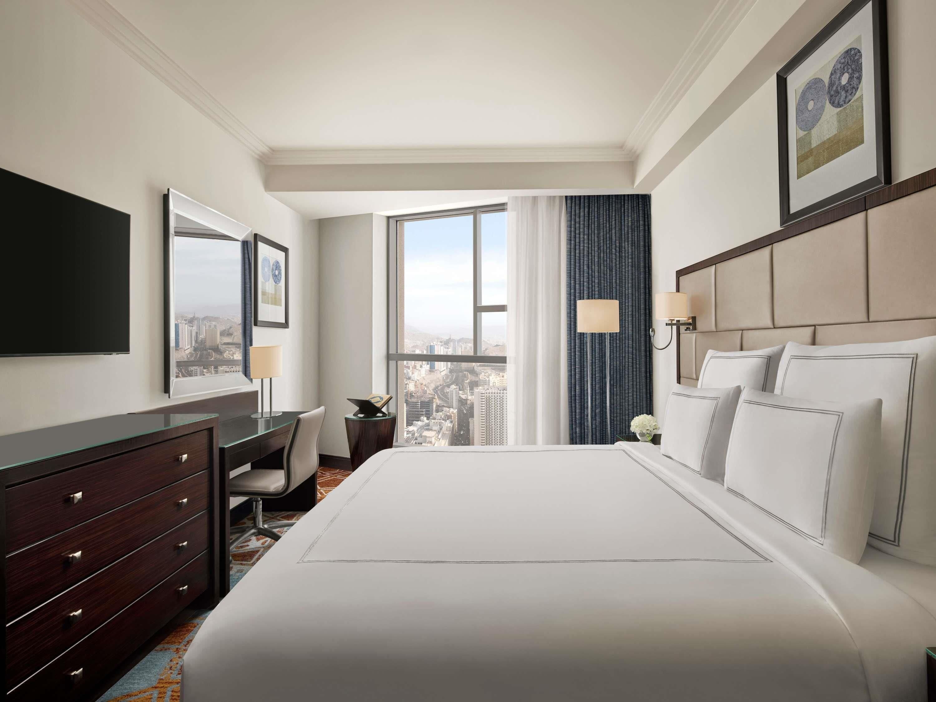 Une chambre d’hôtel avec un grand lit, un bureau, une chaise, une commode, une télévision et une fenêtre donnant sur la ville.