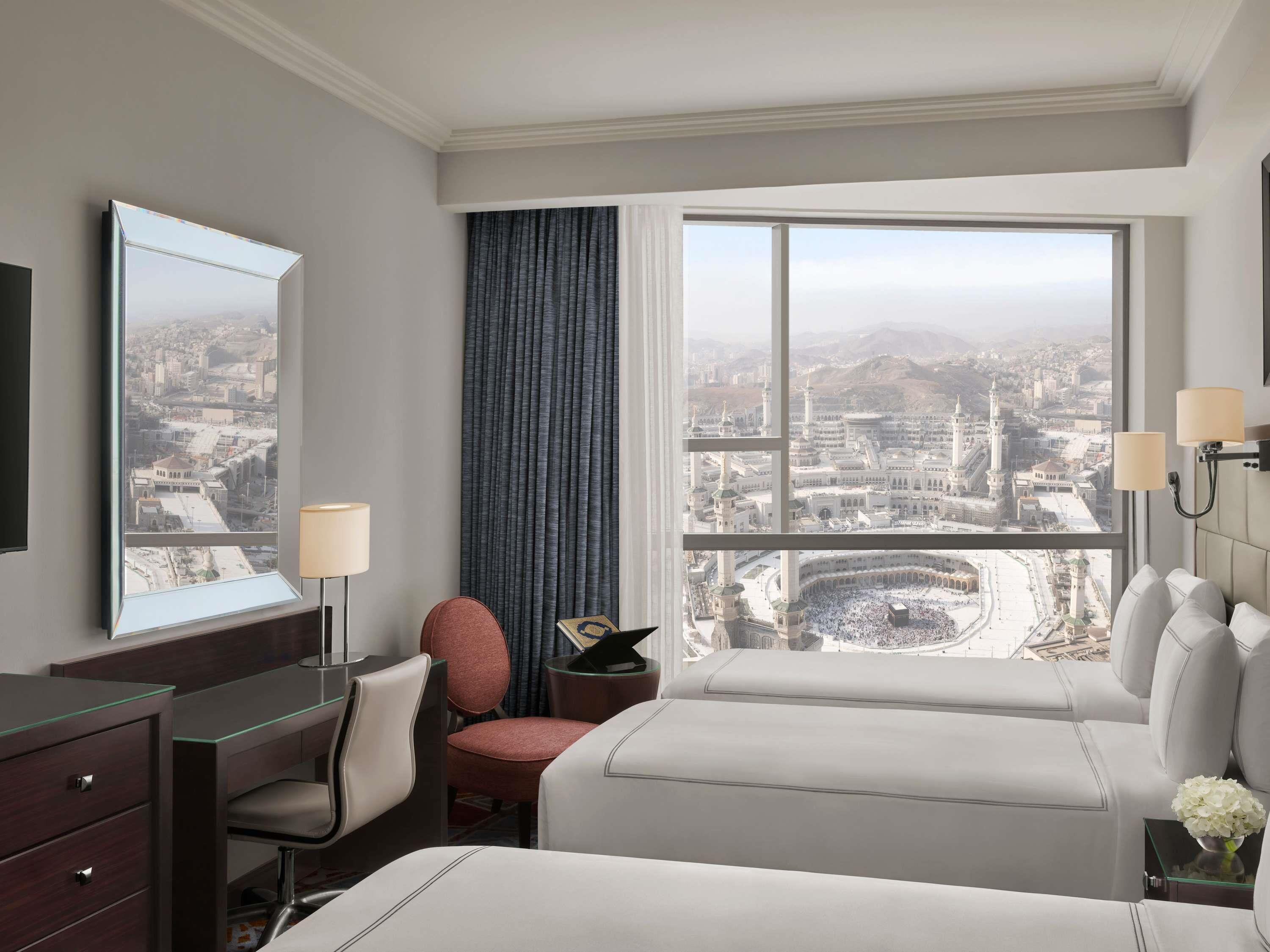 A hotel room with a large window, a desk with a lamp, a chair, a bed, and a nightstand with a vase of flowers.