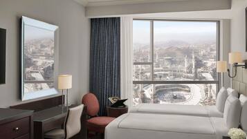 A hotel room with a large window, a desk with a lamp, a chair, a bed, and a nightstand with a vase of flowers.