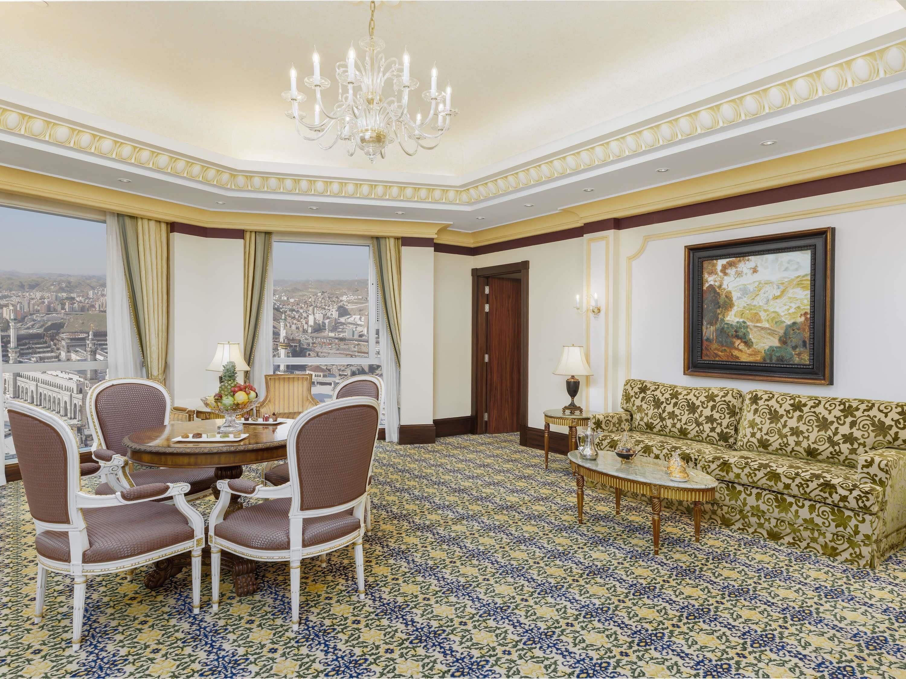 A hotel room with a dining area, a sofa, a chandelier, and a large window offering a city view.