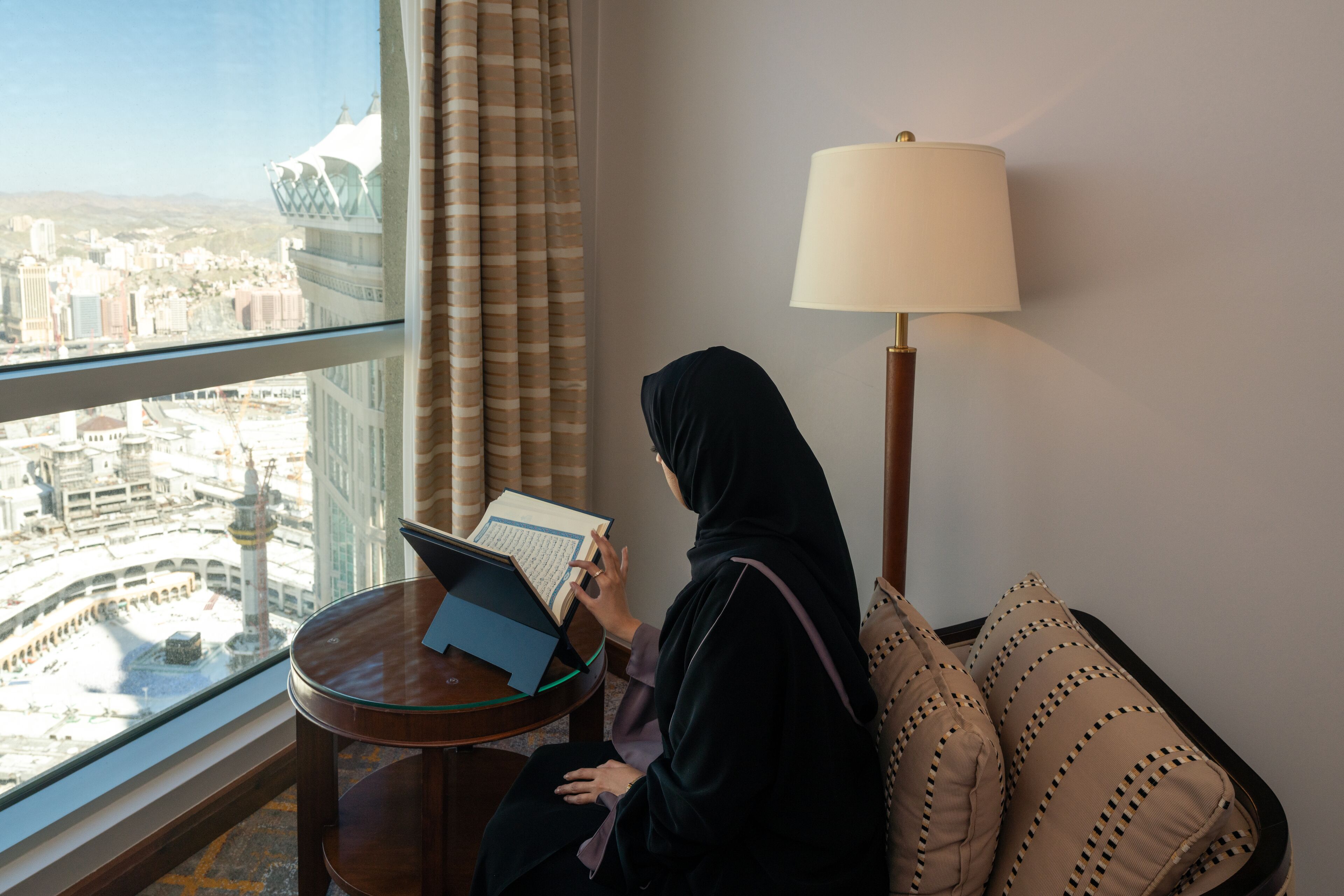 A room with a view of a cityscape, a table with a laptop, a lamp, and a sofa with patterned cushions.