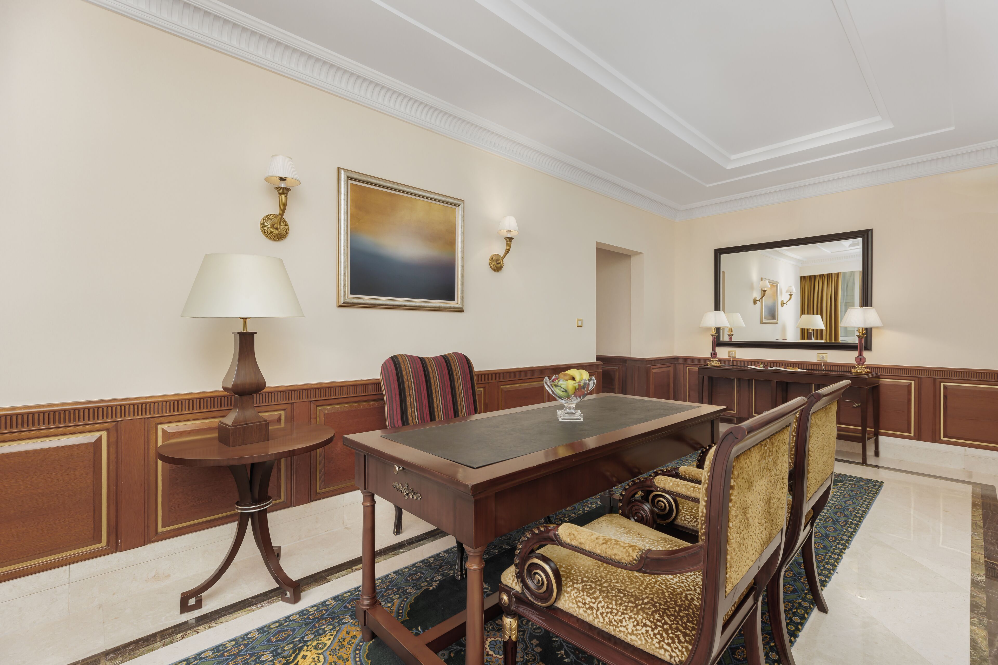 A formal dining room with a wooden table, upholstered chairs, a side table with a lamp, a painting, wall sconces, and a mirror.