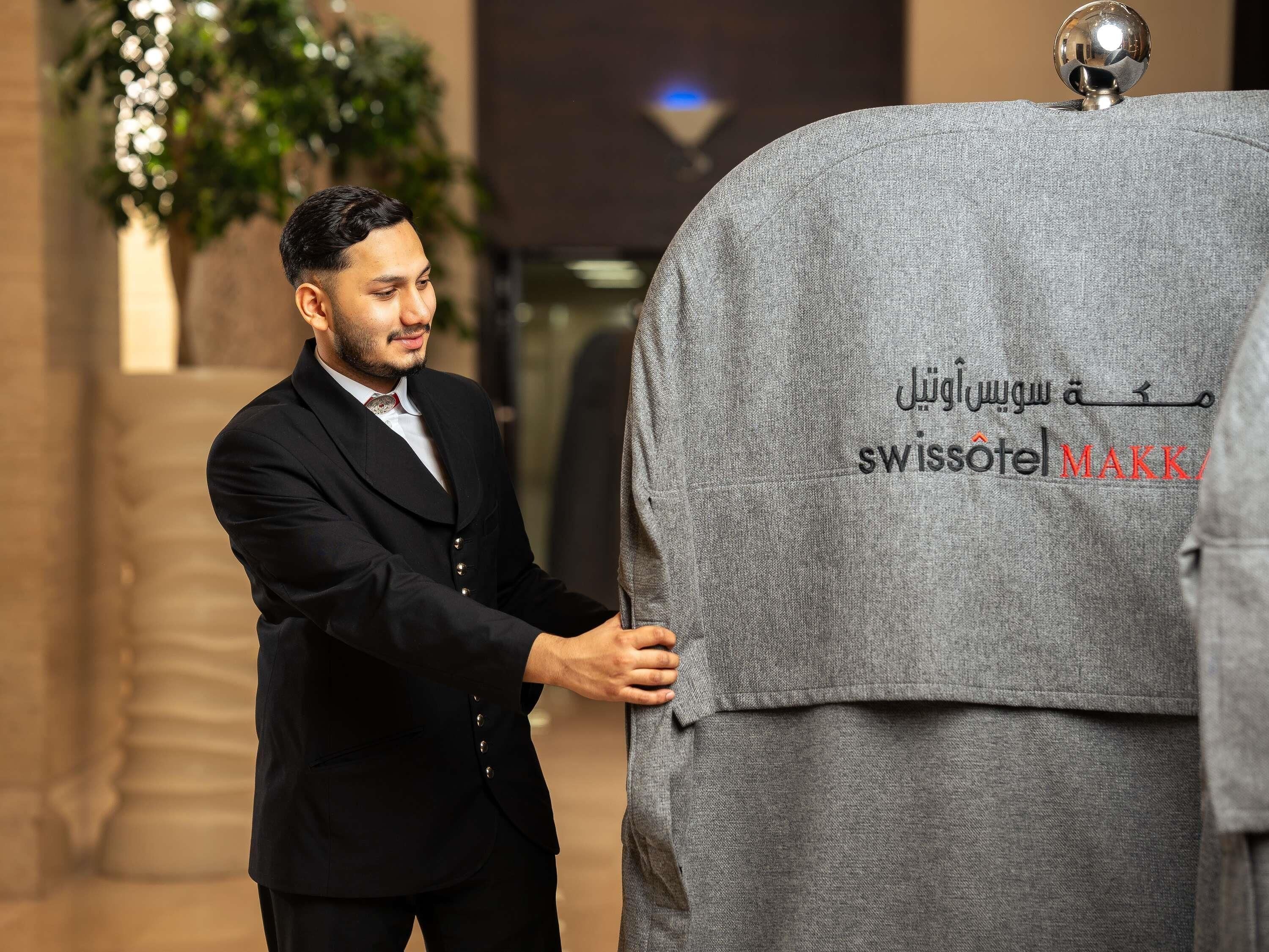 A person in a black suit adjusting a grey hotel garment with Arabic text and 'SWISSOTEL' visible.