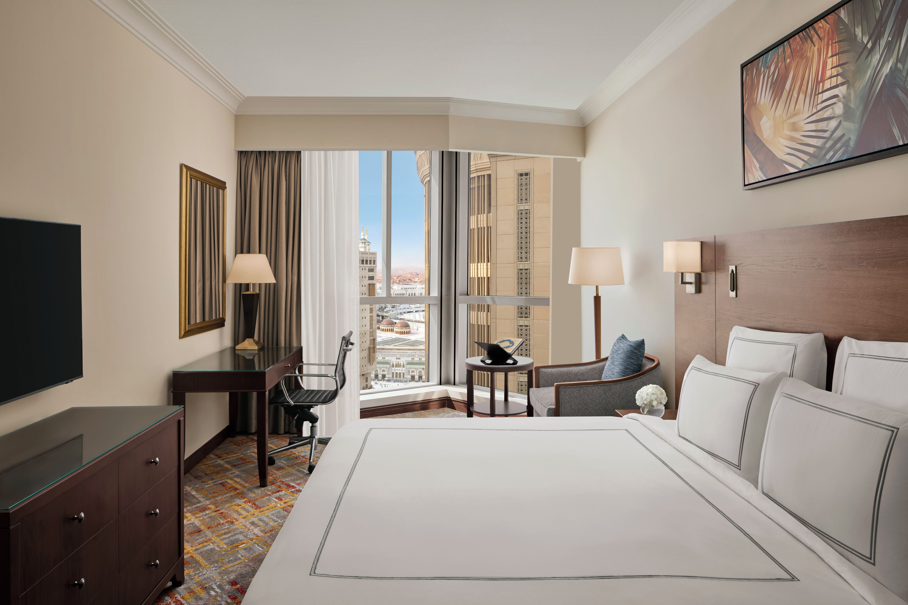 A hotel room with a large bed, a desk, a chair, a TV, and a view of a cityscape through the window.