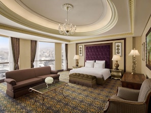 Habitación de hotel con una cama grande, un sofá, una mesa de centro, una silla, una araña y vistas a la ciudad.