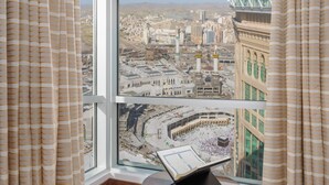 Une pièce dotée d’une grande fenêtre donnant sur un paysage urbain, avec notamment une mosquée et un bâtiment moderne. On y trouve une table ronde en bois sur laquelle est posé un ordinateur portable, ainsi qu’un tapis à motifs sur le sol.