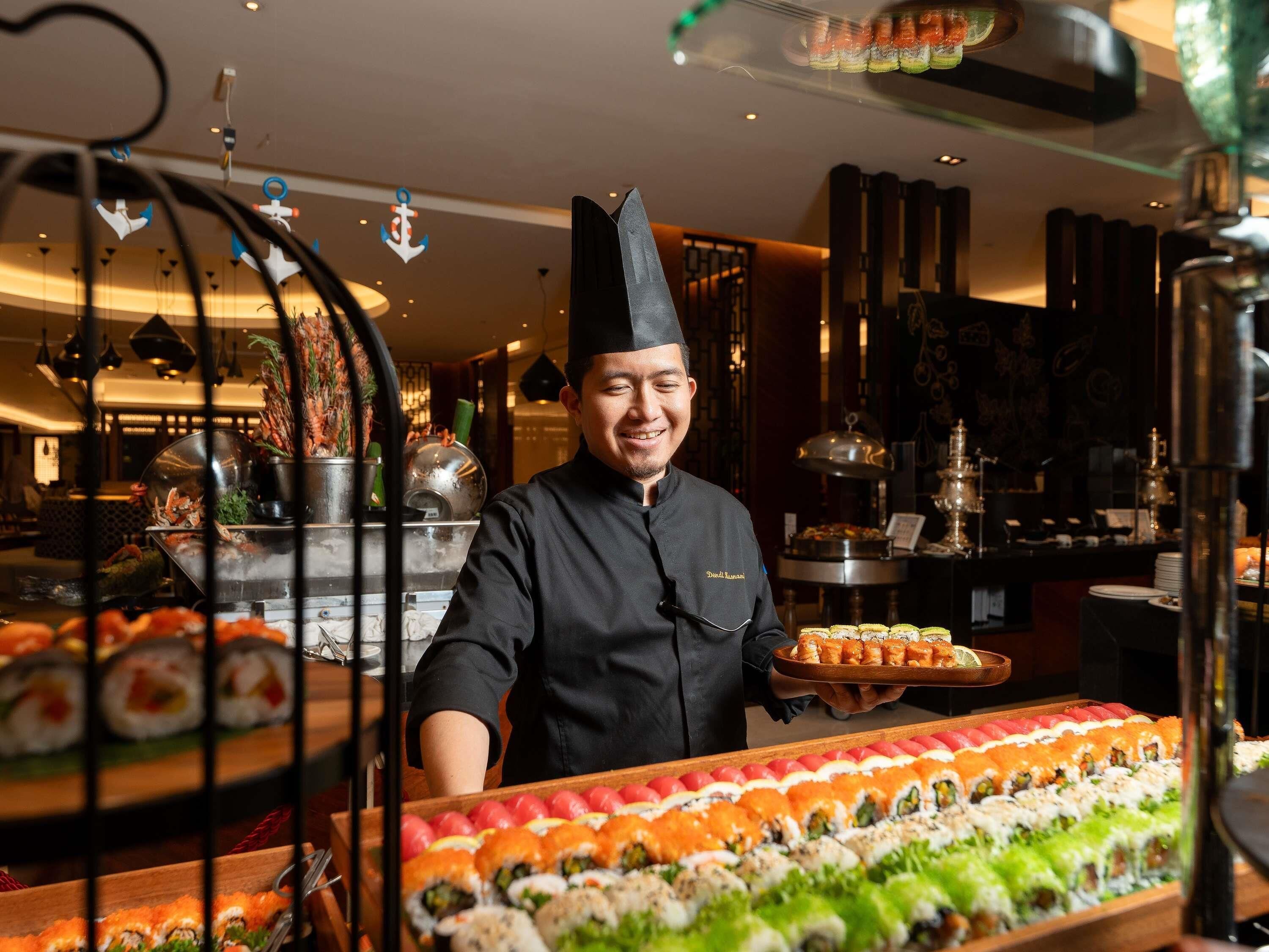 A chef in a black uniform with a name tag, presenting a tray of sushi to a customer.