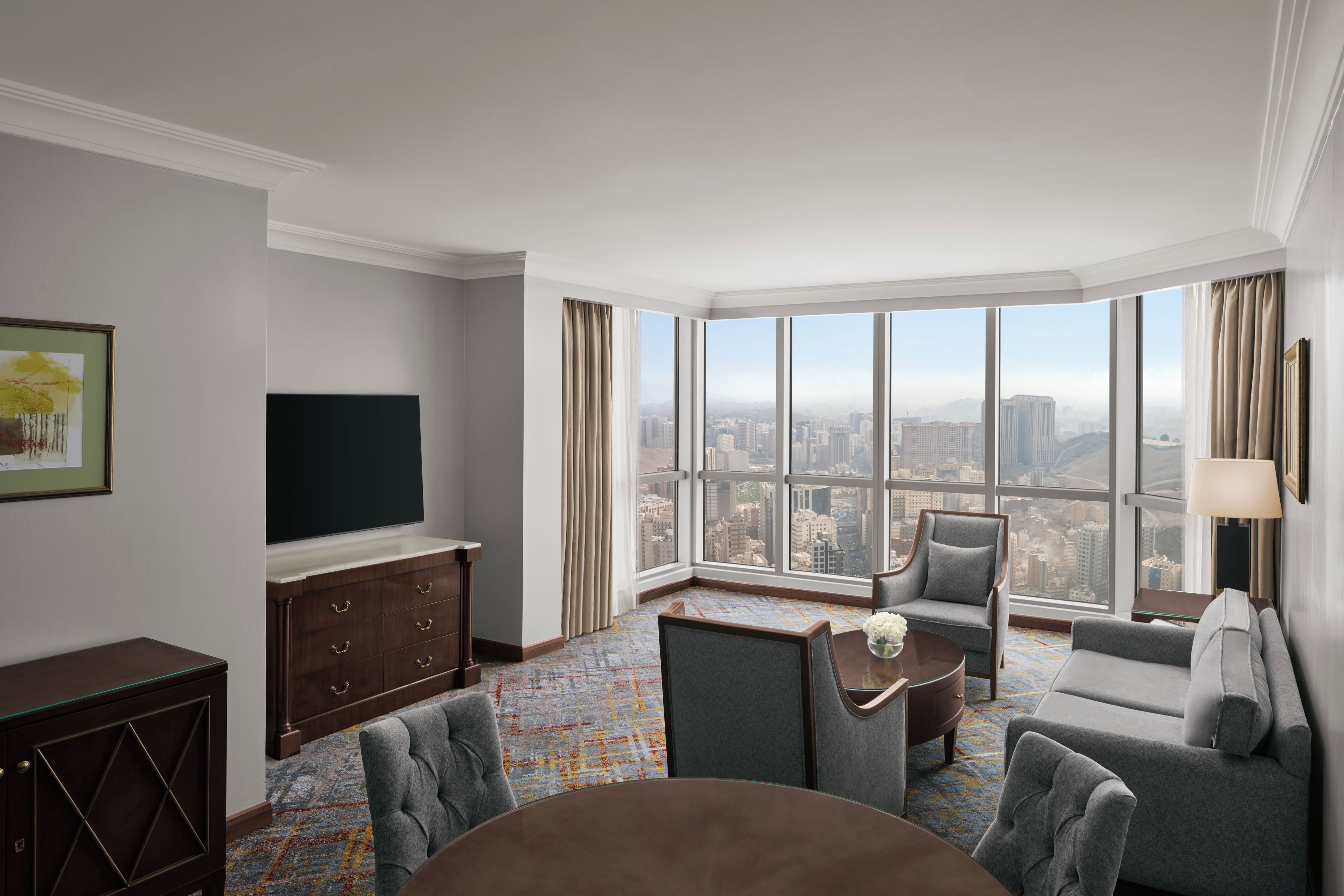 A high-rise hotel room with a large city view, a flat-screen TV, a sofa, a chair, and a small table.