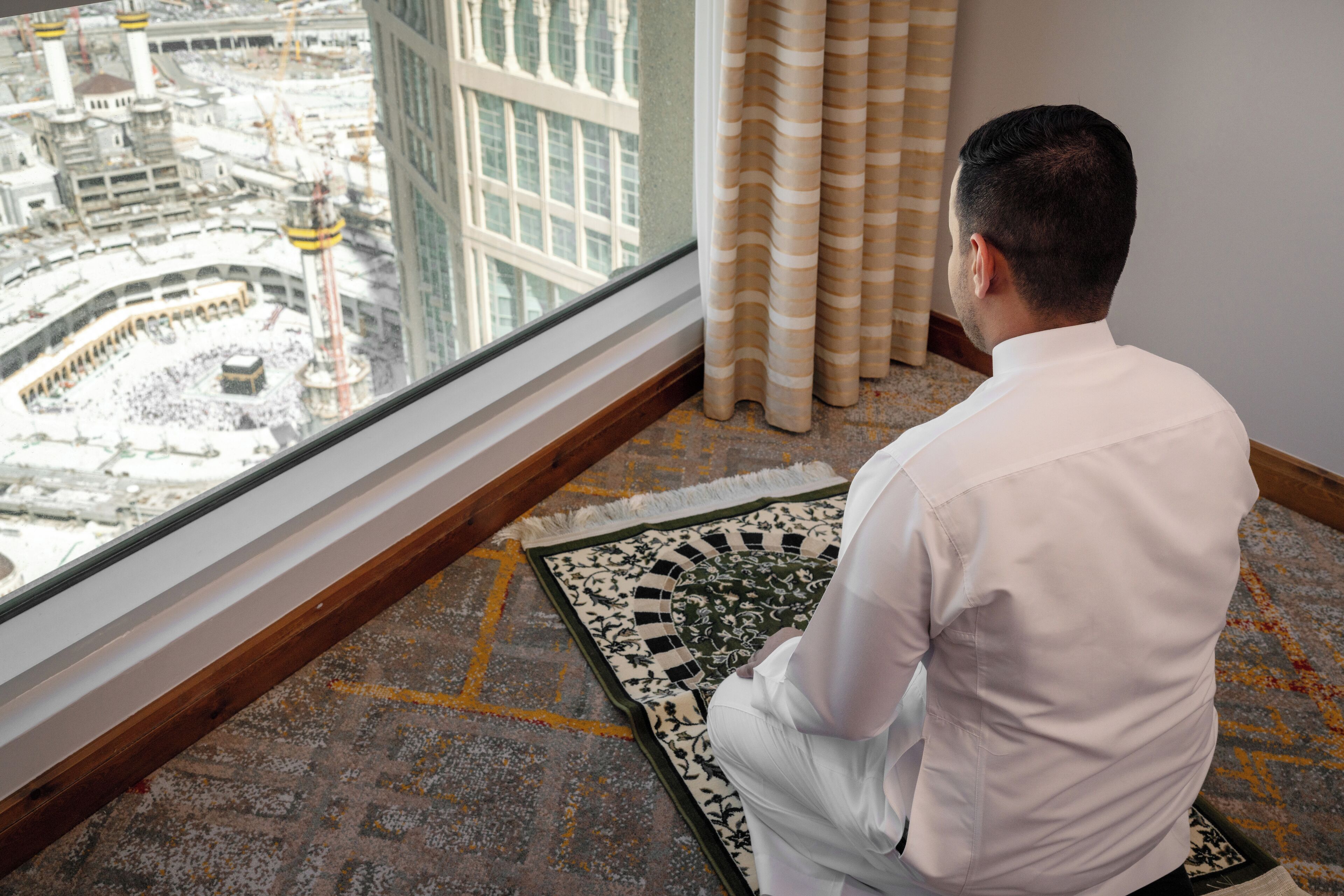 En man sitter på en bönsmatta i ett rum med utsikt över en byggnad med ett minaret.