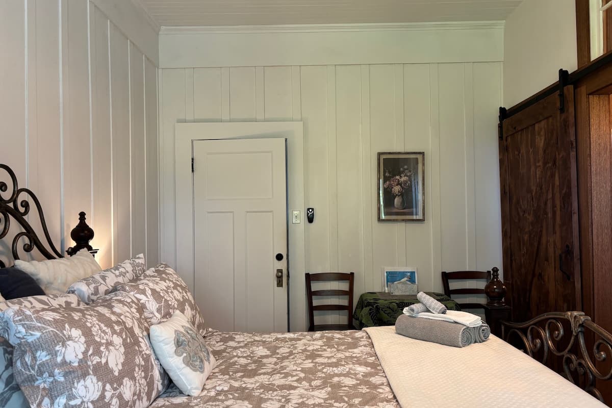 Un dormitorio con cama, dos sillas, un armario de madera, una puerta blanca y un cuadro enmarcado en la pared.
