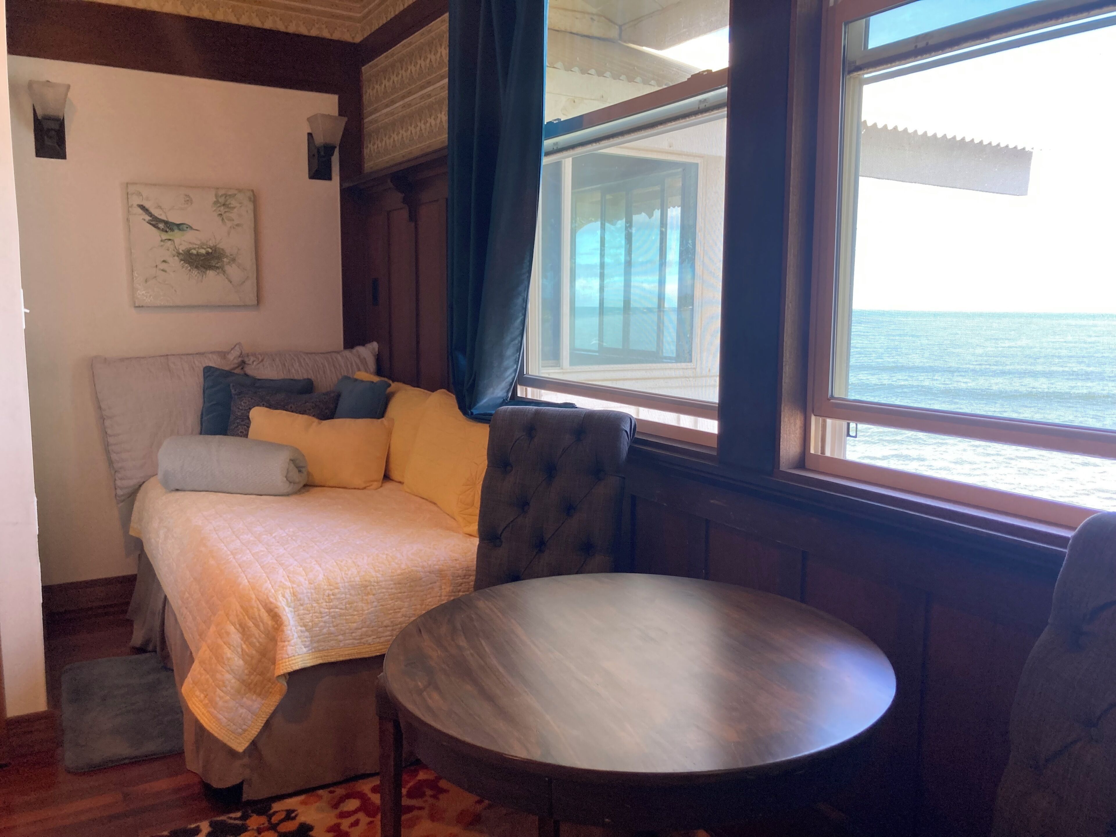 A cozy bedroom with a bed, a round wooden table, and a view of the sea through large windows.