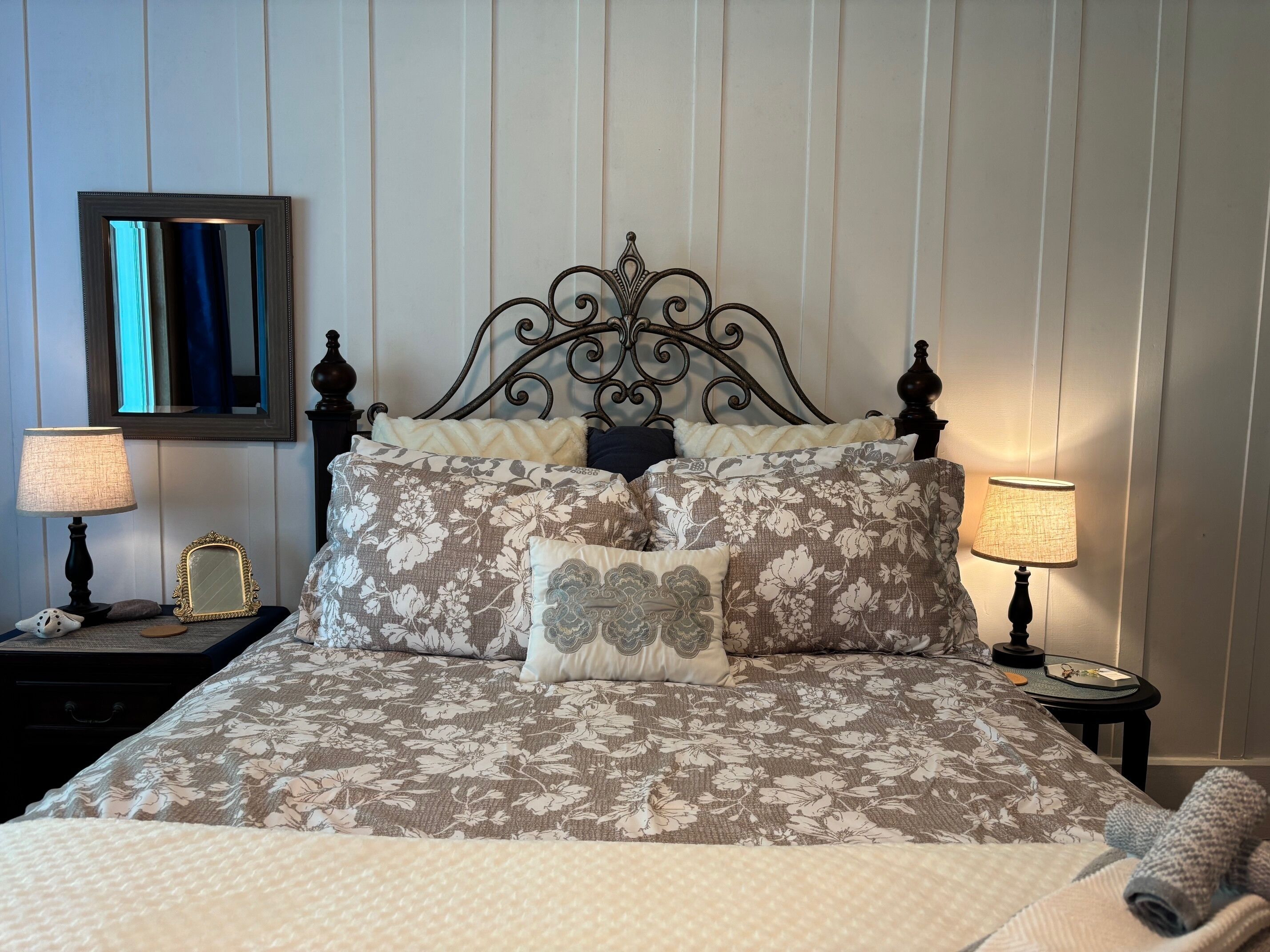 A neatly made bed with floral bedding, two bedside lamps, a mirror, and a small table with a book and a cup.