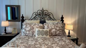 A neatly made bed with floral bedding, two bedside lamps, a mirror, and a small table with a book and a cup.