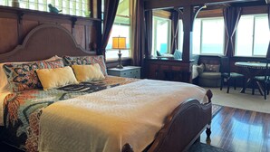 A large bed with a wooden headboard, a nightstand with a lamp, a window with curtains, and a view of the sea. - Hilo Bay Oceanfront B&B (Hilo)