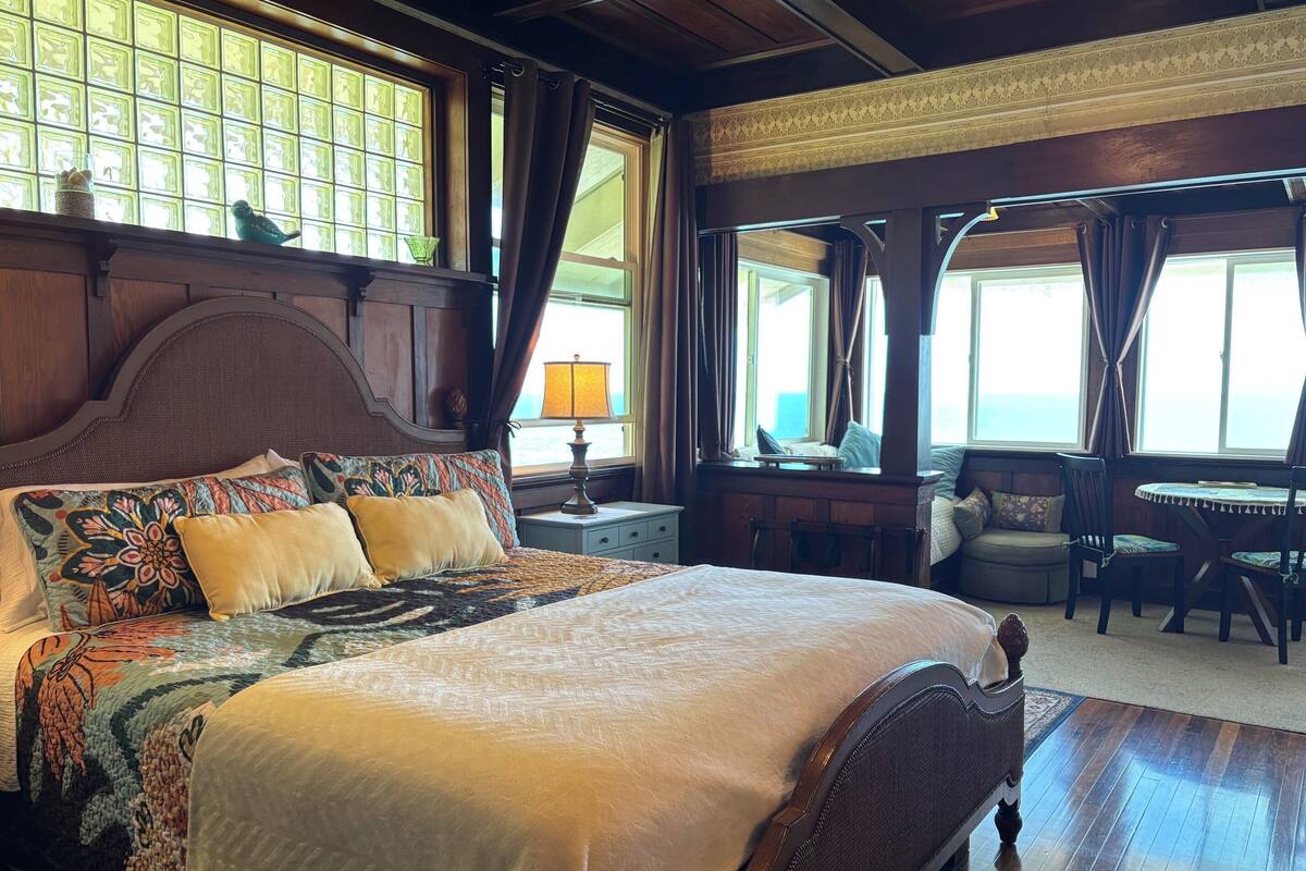 Una cama grande con cabecera de madera, una mesita de noche con lámpara, una ventana con cortinas y vista al mar.
