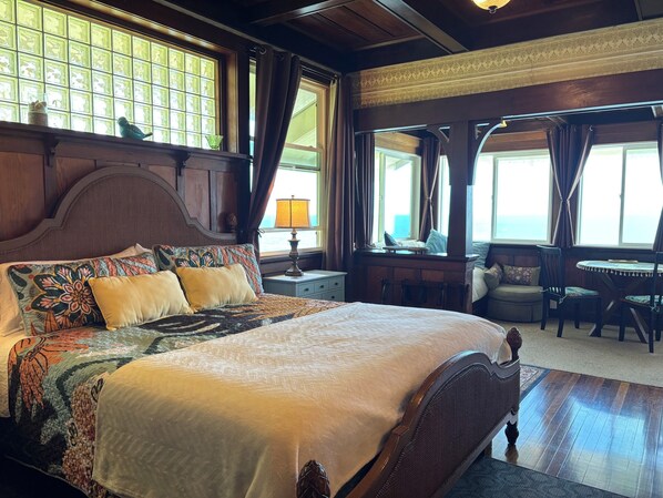 A large bed with a wooden headboard, a nightstand with a lamp, a window with curtains, and a view of the sea. - Hilo Bay Oceanfront B&B (Hilo)