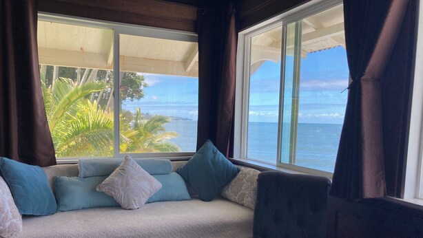 A room with a window overlooking a beach, a sofa with cushions, and a small table. - Hilo Bay Oceanfront B&B (Hilo)