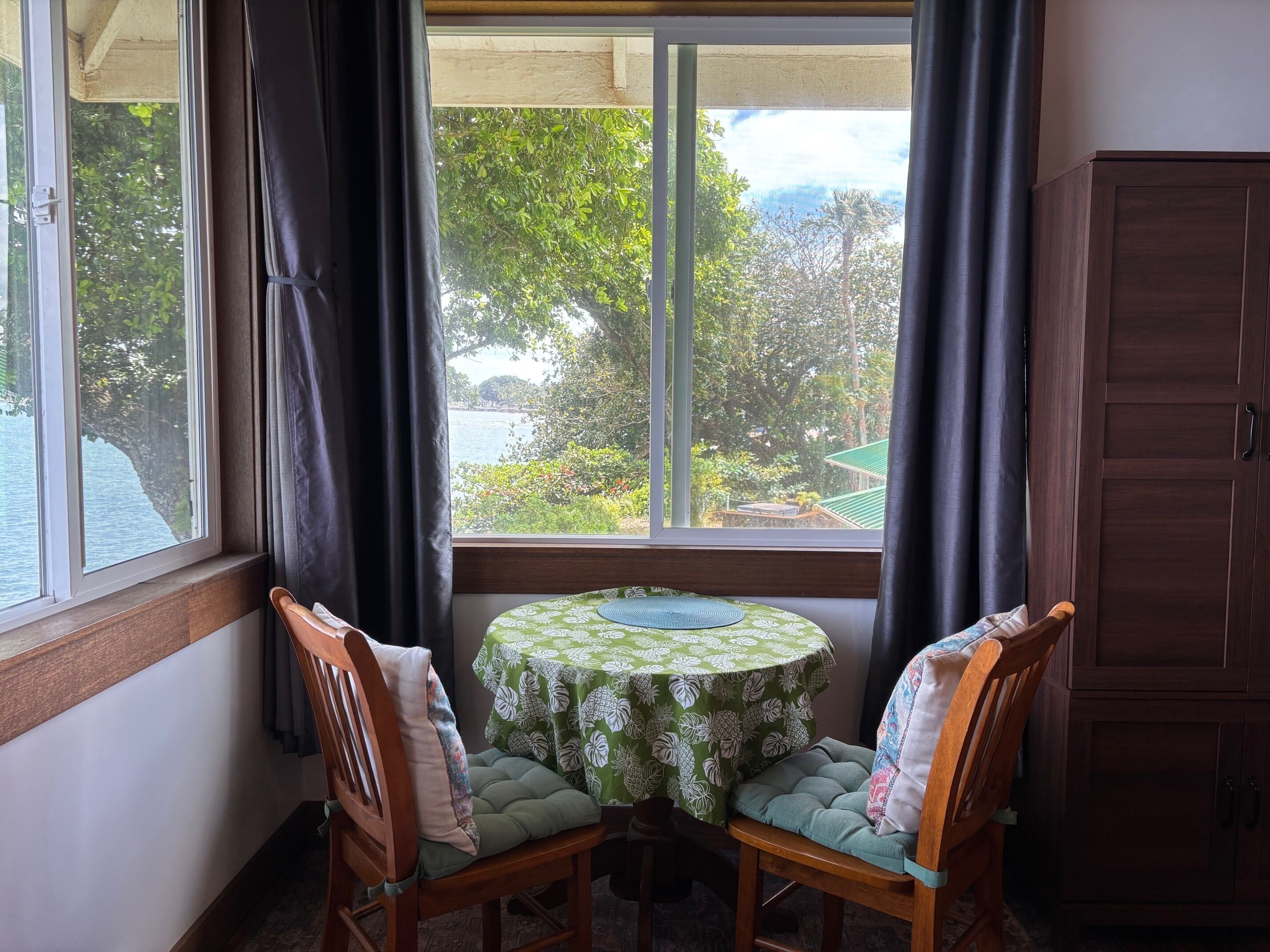 A corner room with a window overlooking a garden and a view of trees and a building. There is a round table with a green floral tablecloth and two wooden chairs with cushions.