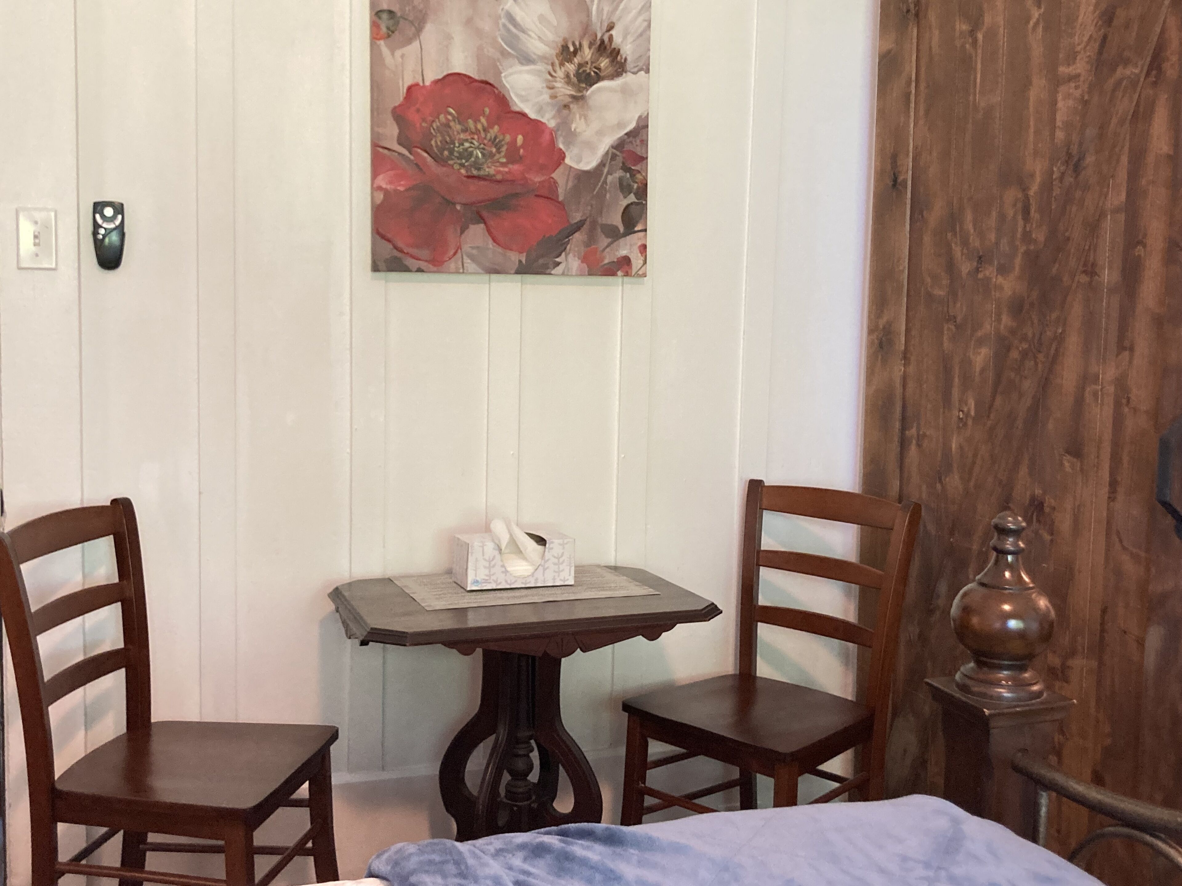 A room with two wooden chairs, a small table with a tissue box, and a bed with a blue blanket.