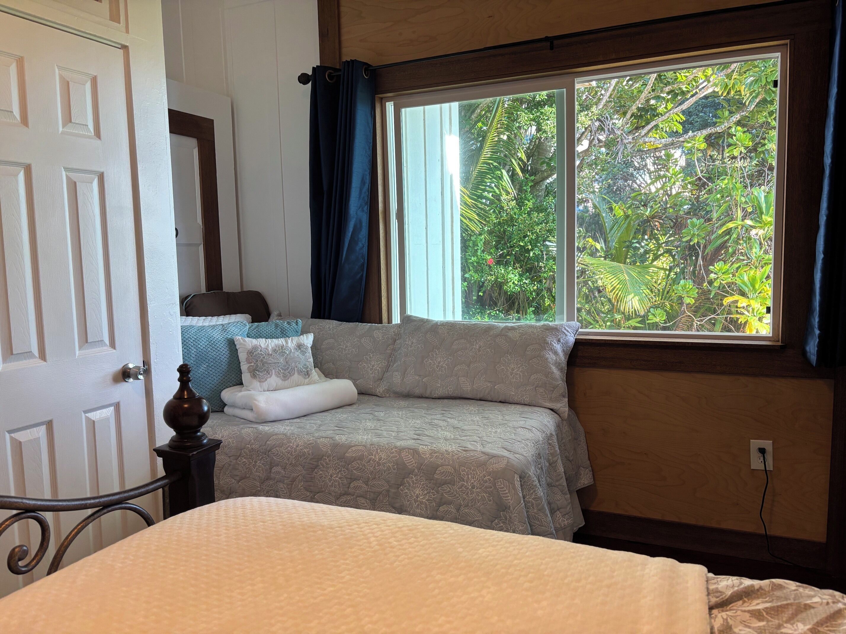 A bedroom with a bed, a window with a view of greenery, a door, and a nightstand.