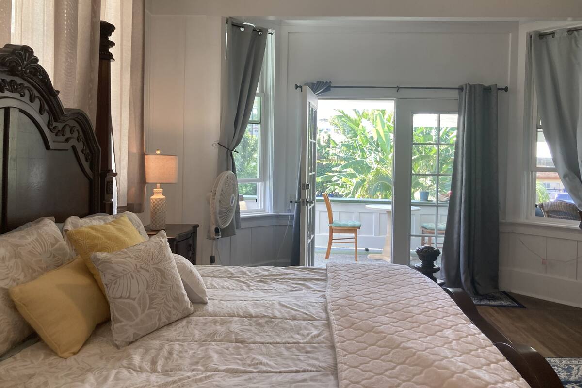 Un dormitorio con una cama grande, una mesita de noche con lámpara, un ventilador y vistas al exterior a través de una puerta.