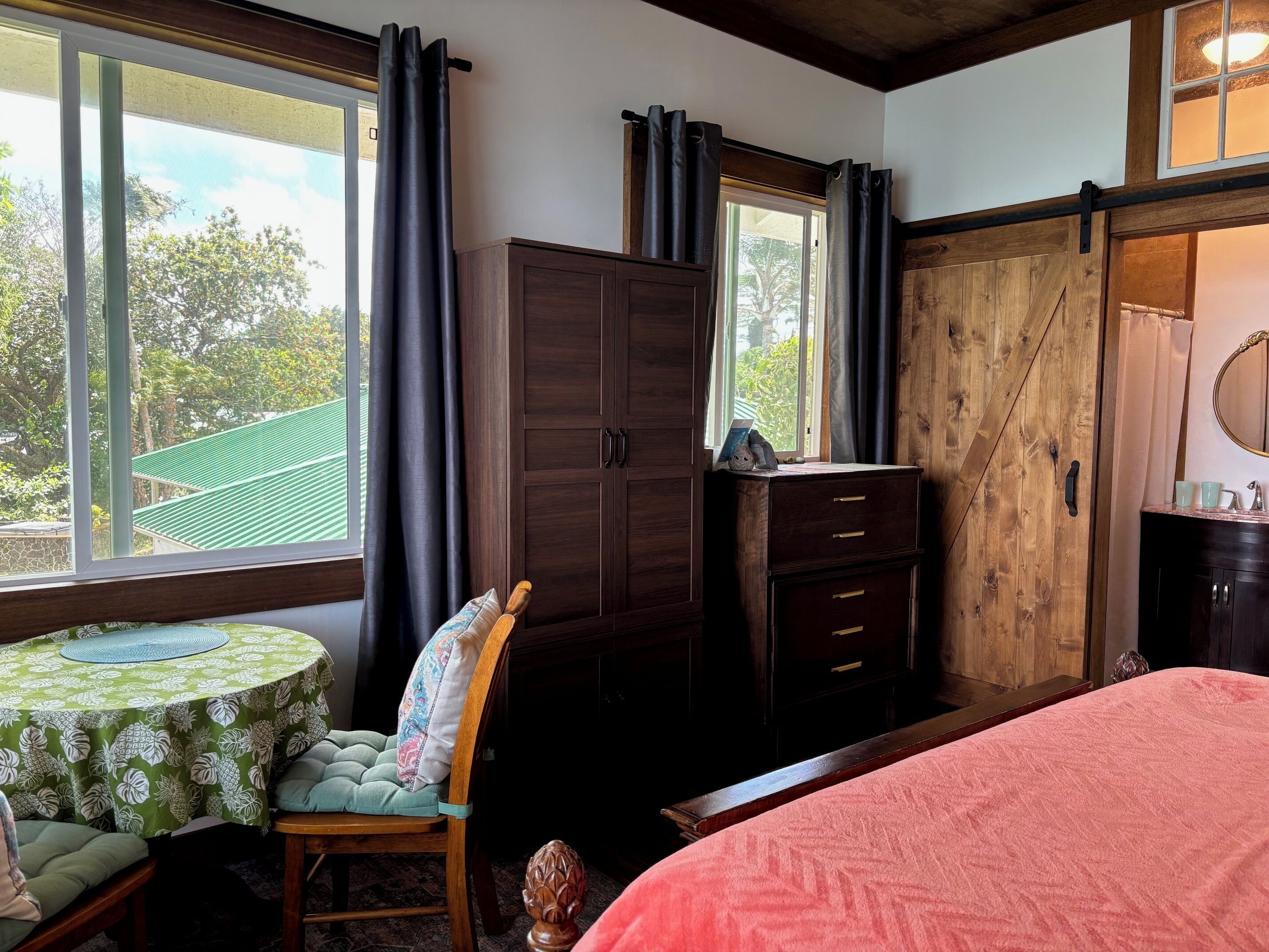 A bedroom with a bed, a wooden wardrobe, a table with a green tablecloth, and a chair with a patterned cushion.