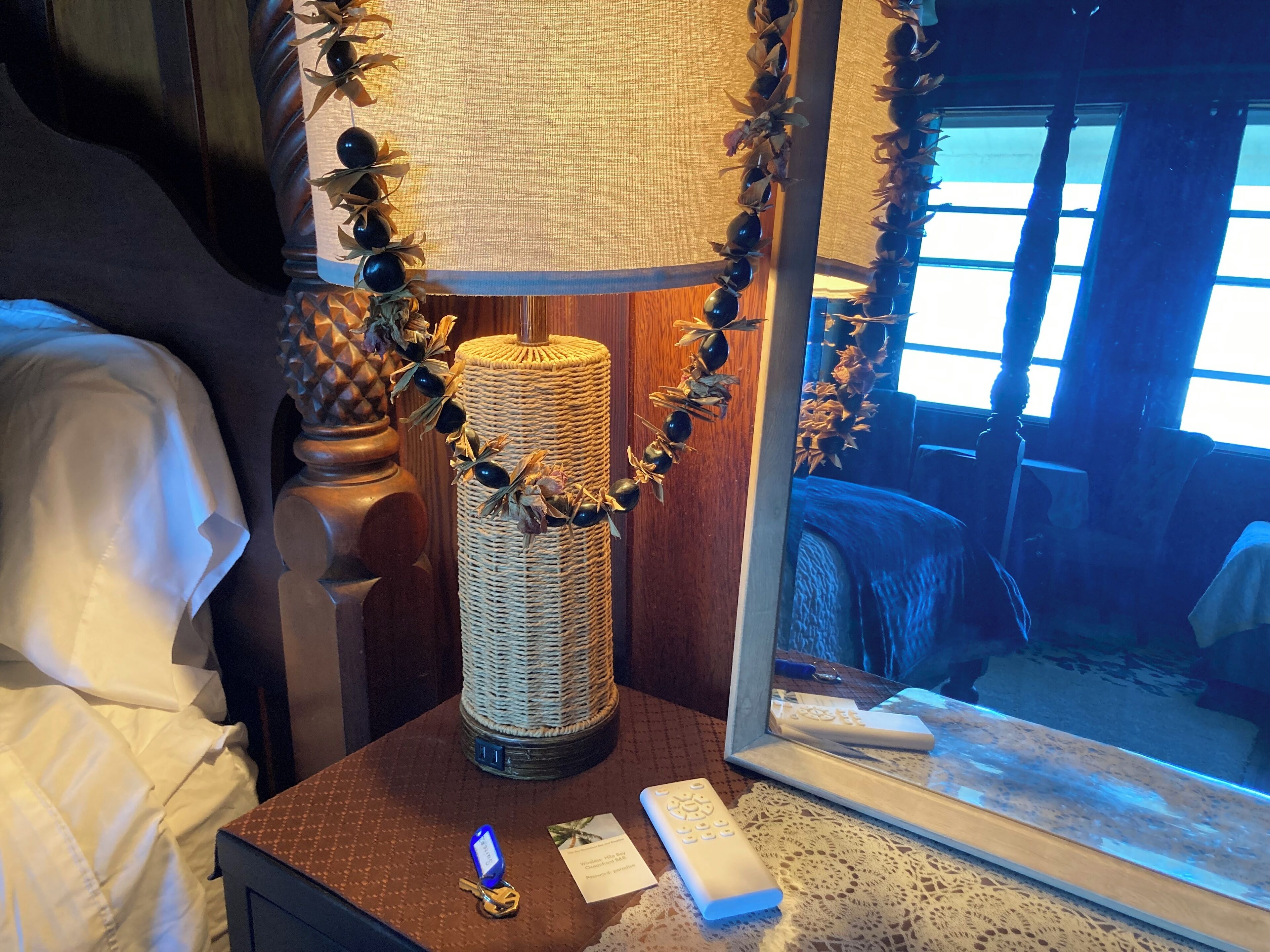 A bed with white linens, a bedside table with a woven lamp, a remote control, and a mirror reflecting a room with a window.