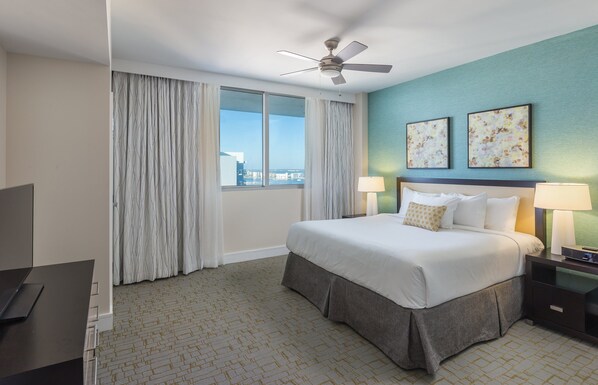 A hotel room with a bed, two bedside lamps, a ceiling fan, a TV, and a view of the city through large windows.