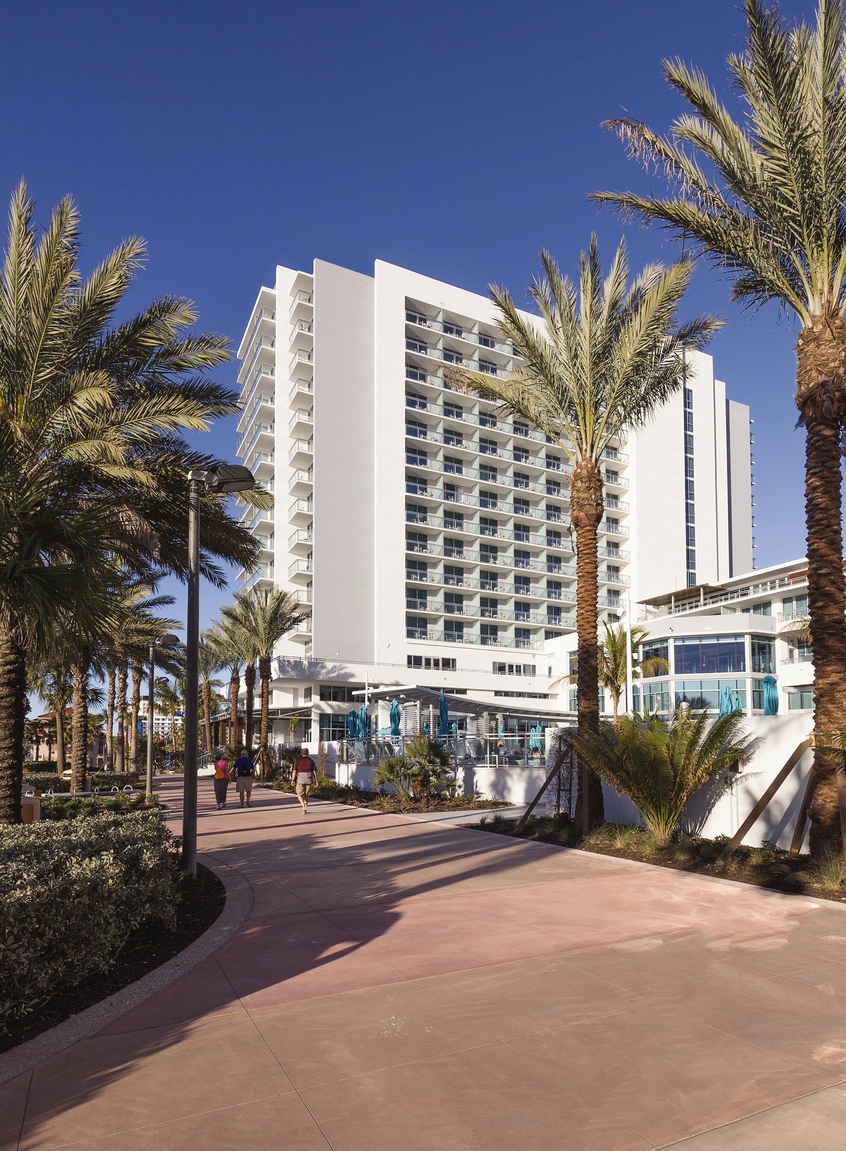 A modern hotel with multiple floors, a covered outdoor area with umbrellas, and palm trees.