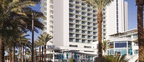 A modern hotel with multiple floors, a covered outdoor area with umbrellas, and palm trees.