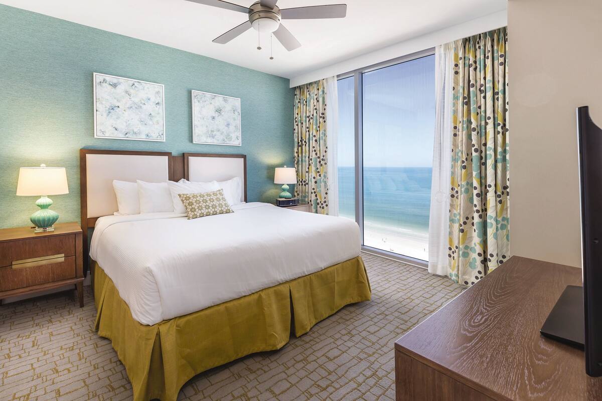 A hotel room with a bed, a desk, a TV, a ceiling fan, and a view of the ocean.