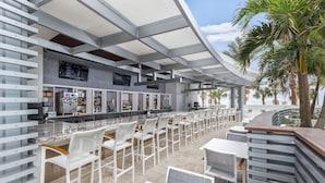 Un área de bar moderna al aire libre con sillas y mesas blancas, una barra con televisor y vistas a palmeras.