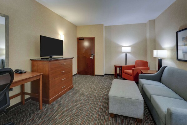 A hotel room with a flat-screen TV on a wooden dresser, a desk with a telephone, a grey sofa, an orange armchair, and a small table with a lamp. - Drury Inn & Suites Charlotte Arrowood (Charlotte)