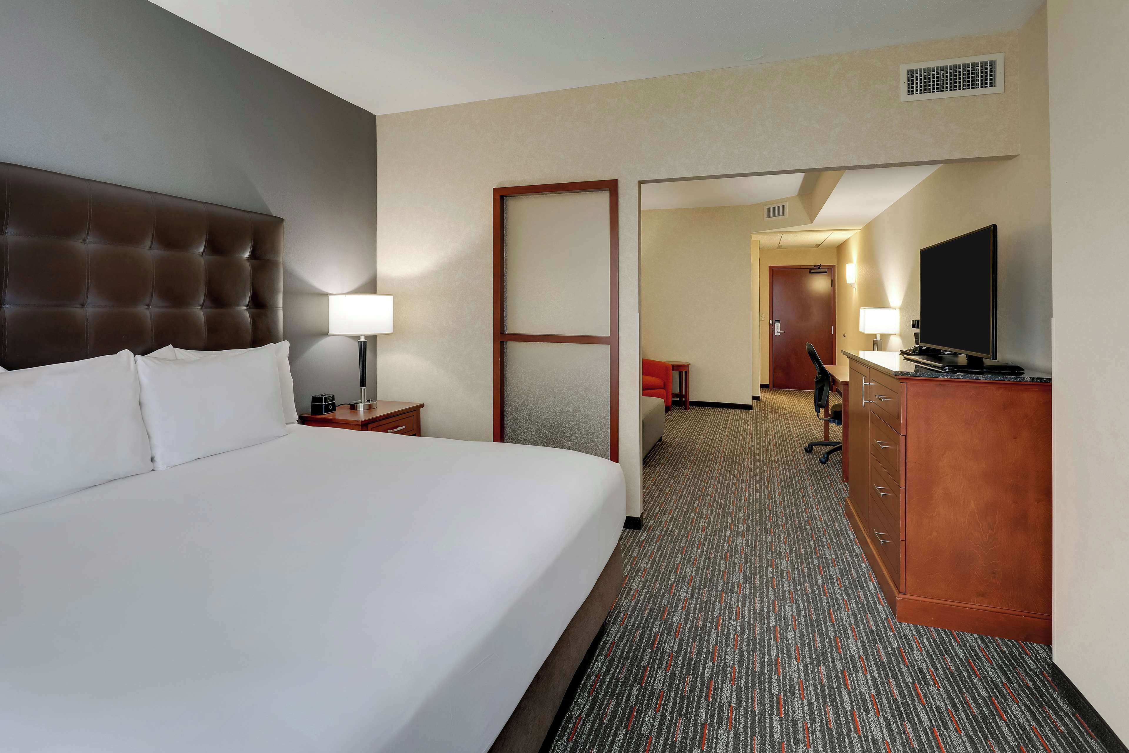 A hotel room with a large bed, a desk with a flat-screen TV, a dresser, and a door leading to a hallway.