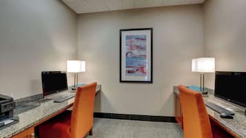 A hotel room with two desks, each with a computer, keyboard, and mouse. There are two lamps on each desk, and a framed abstract painting on the wall.