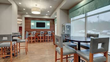 A spacious indoor area with wooden flooring, multiple tables and chairs, a large window with a view of parked cars, and a flat-screen TV mounted on the wall.