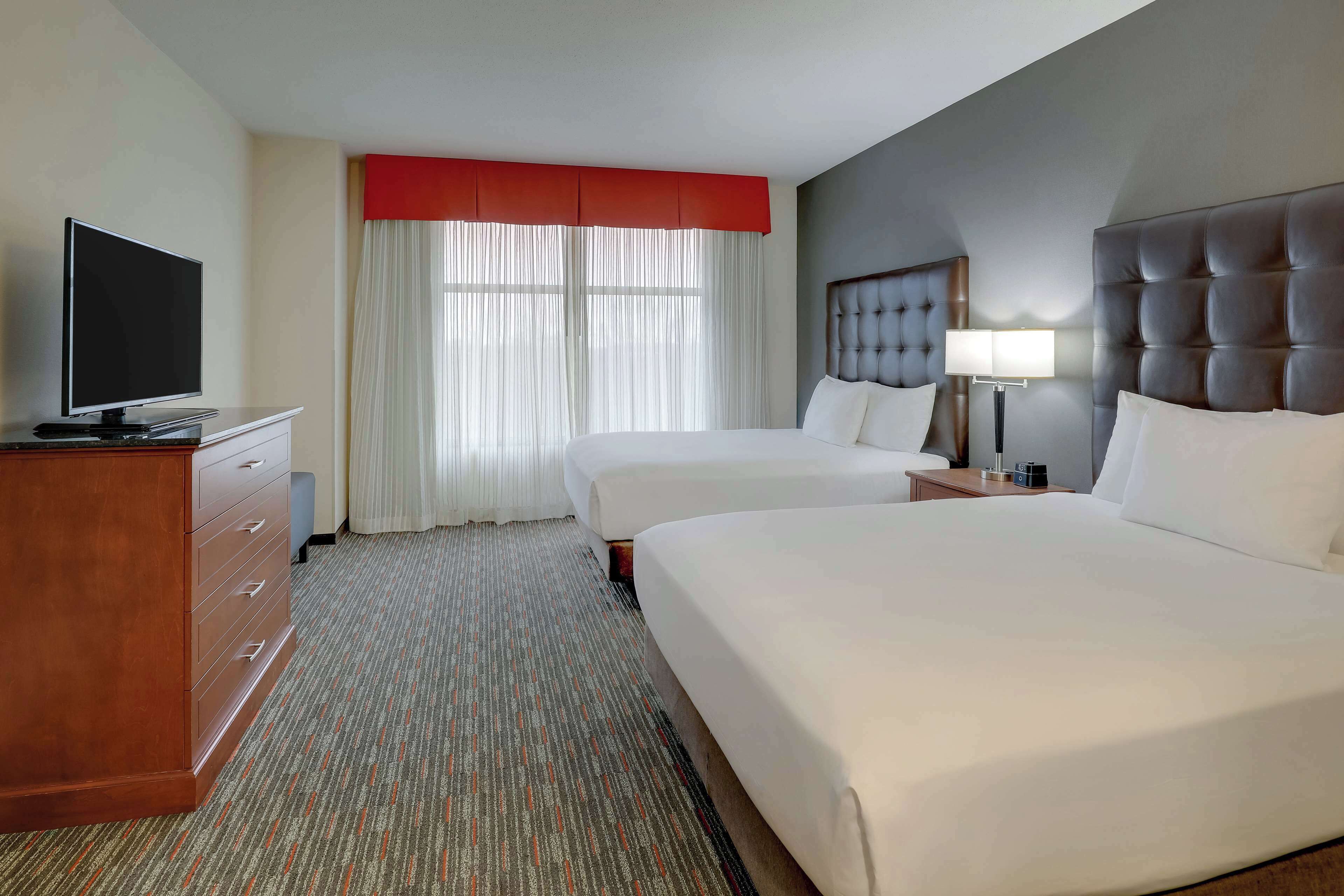 Quarto de hotel com duas camas, televisão, escrivaninha e janela com cortinas.