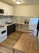 A compact kitchen with a built-in oven, microwave, and refrigerator. There's a wooden dining table and a single chair.
