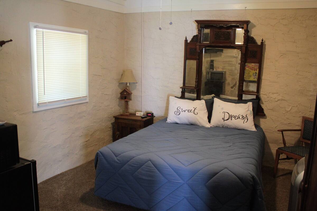 A bedroom with a bed, a nightstand, a lamp, a mirror, and a window with blinds.