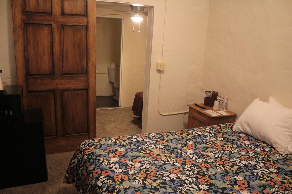 A bedroom with a wooden door, a bed with a floral bedspread, a bedside table with a lamp, and a wall socket.