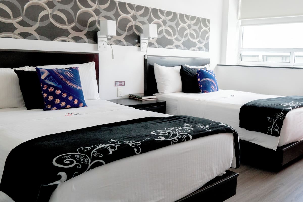 Habitación de hotel con dos camas, cada una con una colcha de diseño blanco y negro, un cabecero y una mesita de noche con lámpara.