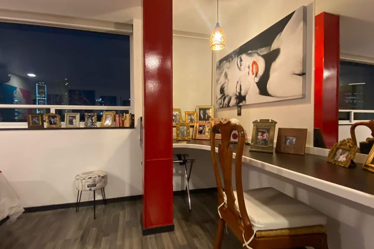 Un dormitorio con un ventanal grande, una cama, un escritorio con silla y una pared decorada con cuadros.