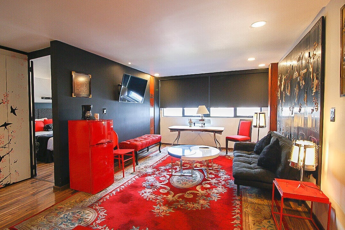 Un espacio habitable compacto con un sofá rojo, un armario rojo, una mesa de centro redonda y una alfombra estampada.